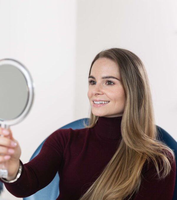 Eine junge Patientin sitzt auf dem Behandlungsstuhl und begutachtet ihr Lächeln in einem Handspiegel.