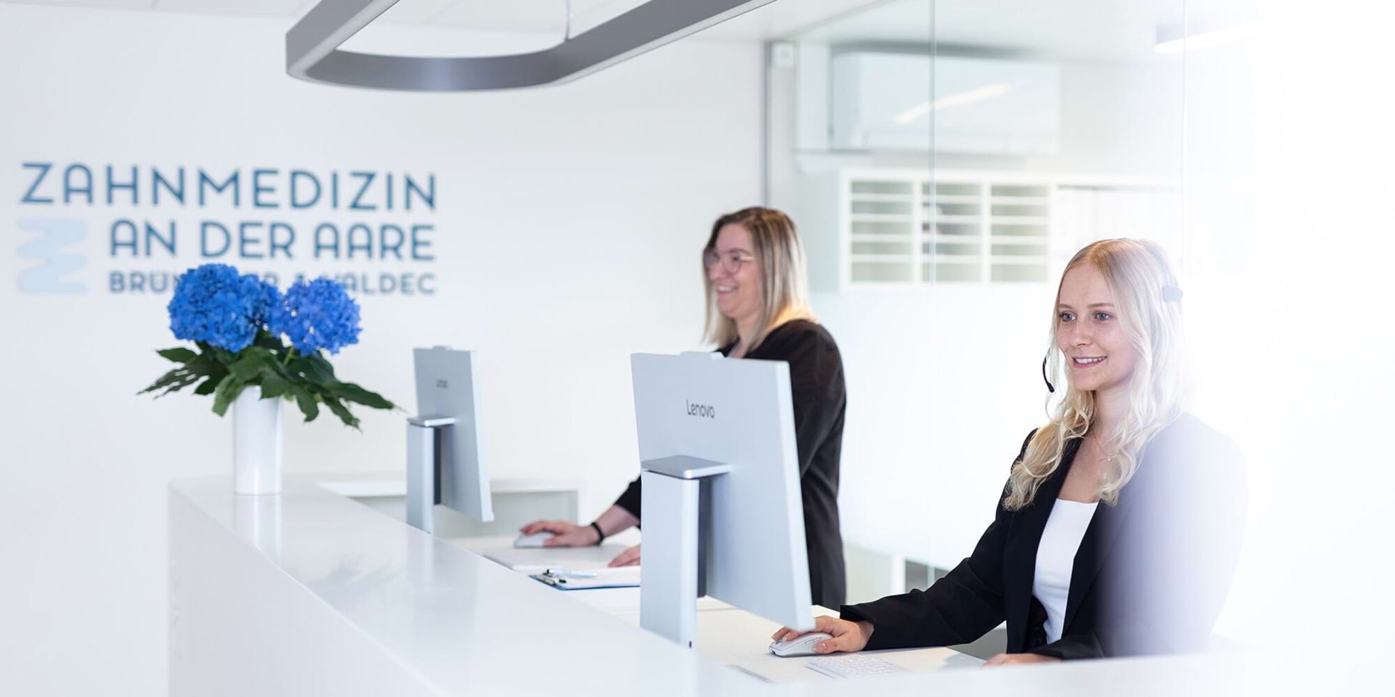 Patientin kommt am Empfang der Praxis an und wird von einer Mitarbeiterin lächelnd begrüsst.