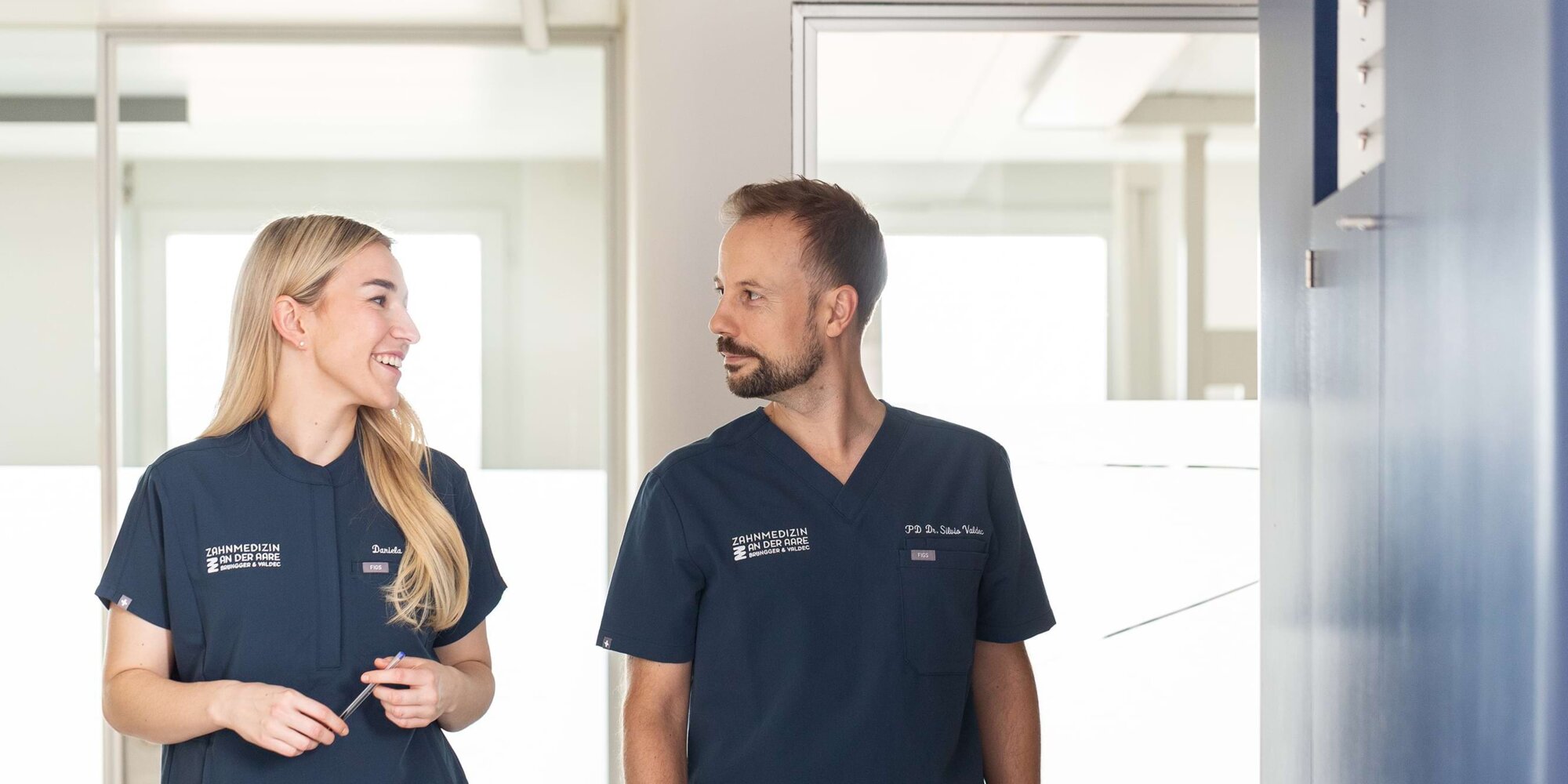 Dr. Daniela Br&uuml;ngger (links) und Dr. Silvio Valdec (rechts) unterhalten sich und laufen vor beleuchtetem Hintergrund durch den Flur der Praxis.