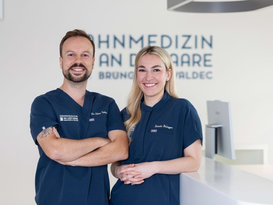 PD Dr. Silvio Valdec (links) und Dr. Daniela Brüngger (rechts) stehen zusammen am Empfang. Im Hintergrund ist das Logo Zahnmedizin an der Aare zu sehen.