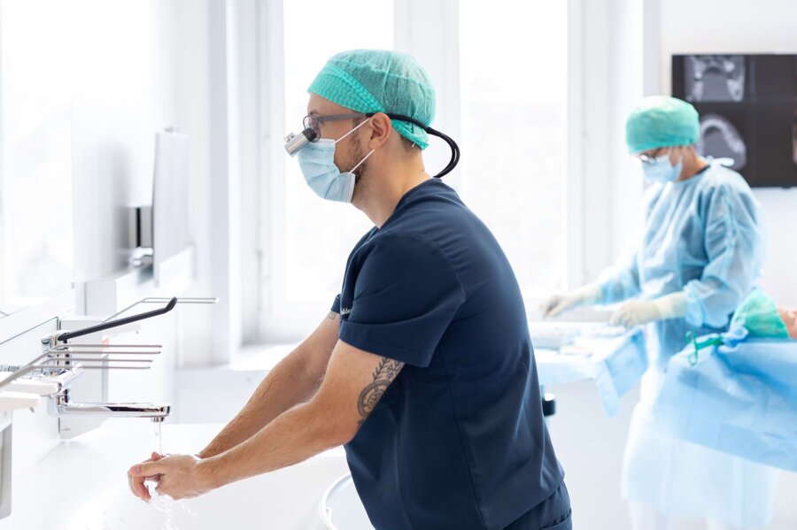 PD Dr. Silvio Valdec bei der Vorbereitung einer OP. Im Hintergrund sind eine Assistenz und der Patient auf der Liege zu sehen.