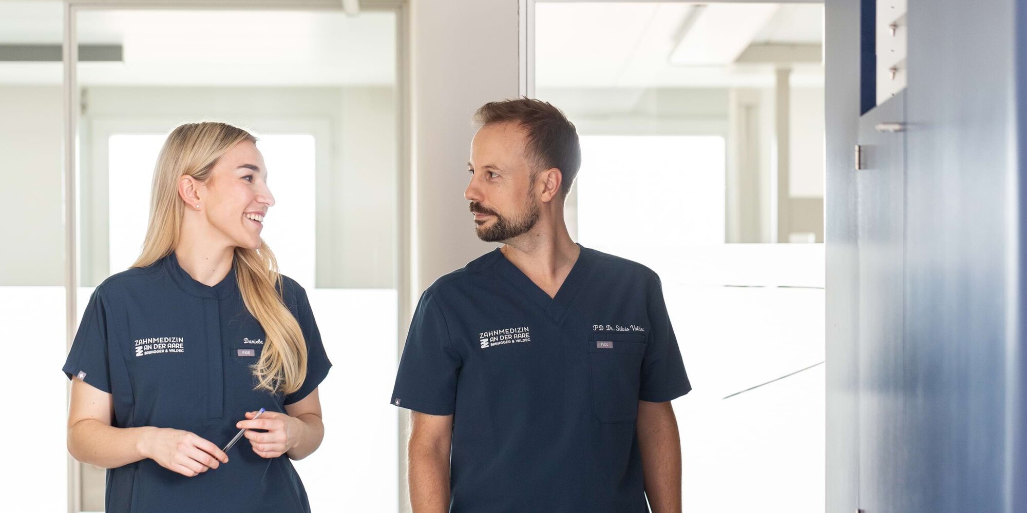 Dr. Daniela Brüngger (links) und Dr. Silvio Valdec (rechts) unterhalten sich und laufen vor beleuchtetem Hintergrund durch den Flur der Praxis.