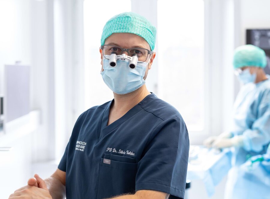 Nahaufnahme von Dr. Silvio Valdec bei der OP-Vorbereitung. Im Hintergrund sind verschwommen eine OP-Assistenz sowie ein Patient auf der Liege zu sehen.